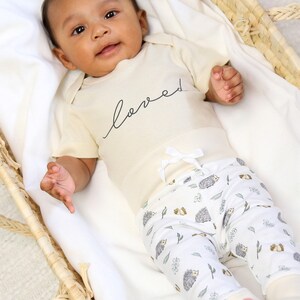 May include: A baby wearing a cream-colored bodysuit with the word "loved" written in black script. The baby is wearing white and cream-colored pants with a hedgehog print. The baby is lying in a wicker basket.