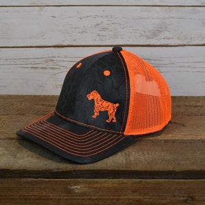 May include: A black and orange camouflage trucker hat with an orange dog silhouette embroidered on the front.