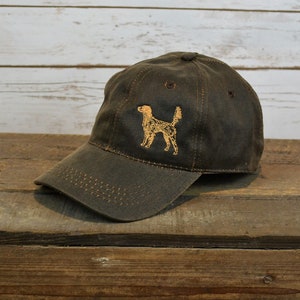 Może przedstawiać: Czapka bejsbolowa z ciemnozielonej tkaniny z haftowanym pomarańczowym sylwetem psa z przodu.