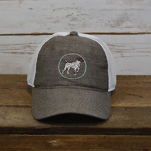 May include: A gray and white trucker hat with a brown and white dog embroidered on the front. The dog is wearing a light blue collar and is standing inside a light green circle.