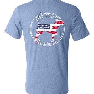 May include: A light blue t-shirt with a white circle graphic on the back. The graphic features a silhouette of a dog with an American flag pattern and the text "Hunt Dog Mania Hunt the USA".