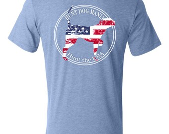 May include: A light blue t-shirt with a white circle graphic on the back. The graphic features a silhouette of a dog with an American flag pattern and the text "Hunt Dog Mania Hunt the USA".
