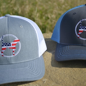 May include: Two gray and blue trucker hats with a white mesh back. Each hat features a white embroidered circle with a silhouette of a dog with an American flag design.