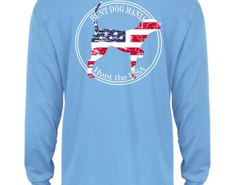 May include: Light blue long-sleeve shirt with a white circle graphic on the back. The graphic features a silhouette of a dog with an American flag pattern and the text "Hunt Dog Mania Hunt the USA".