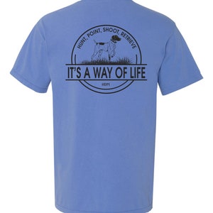 May include: A light blue t-shirt with a black graphic on the back. The graphic features a hunting dog, a circle with the text "Hunt, Point, Shoot, Retrieve" and the text "It's a way of life".