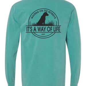 May include: Teal long-sleeve t-shirt with a black graphic of a dog sitting in grass with the text "Waiting on the Retrieve. It's a way of life. HDM" in a circle.