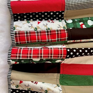 May include: A patchwork Christmas-themed quilt with various fabric patterns. The quilt features sections of red polka dots, plaid, reindeer, snowmen, and black and white patterns. The edge is trimmed with a black and white striped fabric. The word "Shine" is visible.
