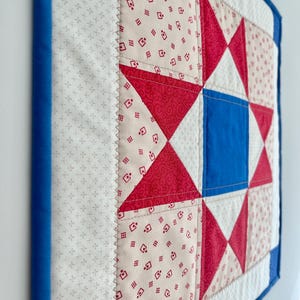 May include: A quilted wall hanging featuring a star pattern in red, white, and blue. The star is centered on a white background with a blue border. The quilt is made of fabric with a repeating pattern.
