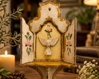 May include: A wall-mounted wooden shrine with a distressed cream and gold finish. The central panel features a carved white dove and a painted pink rose. The two side panels are decorated with painted green leaves and pink roses on a cream background.