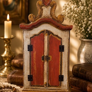 Puede incluir: Un relicario rectangular de madera con un acabado desgastado en blanco y rojo, que presenta dos puertas con bisagras y herrajes de metal negro. La estructura tiene una parte superior arqueada con volutas decorativas y una base plana.