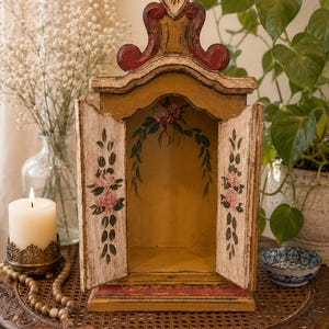 Puede incluir: Un santuario de madera con dos puertas abiertas, pintado en un acabado amarillo mostaza con bordes desgastados. El interior y las puertas presentan motivos florales pintados a mano en rosa y verde. La parte superior tiene un frontón arqueado.