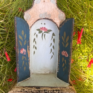 Puede incluir: Un pequeño santuario de madera con parte superior arqueada, que presenta dos puertas abatibles pintadas en azul oscuro con motivos florales. El panel interior es azul claro con una flor rosa y enredaderas verdes pintadas. Los bordes tienen un acabado desgastado.