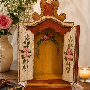 Puede incluir: Un santuario de madera con parte superior arqueada, pintado en rojo y amarillo con un acabado desgastado. Las dos puertas abiertas presentan rosas rosas y hojas verdes pintadas a mano, mientras el panel interior muestra una guirnalda floral.
