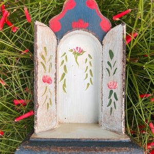 May include: A small wooden shrine with an arched top, featuring a dark blue and red painted crest. The interior is off-white with hand-painted green vines and pink floral accents on the back panel and two side doors, set on a dark base.
