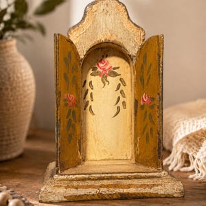 May include: A decorative wooden shrine with a distressed mustard yellow and cream finish. The piece features an arched top, two side panels, and a central alcove hand-painted with pink rose motifs and green foliage accents on a cream background.