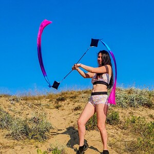 May include: A person in a pink and blue bikini top and matching shorts is twirling a long, flowing ribbon in the air. The ribbon is attached to a black pole and is flowing in the wind. The person is standing on a sandy hill with green grass and plants in the background. The sky is blue.