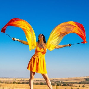Könnte beinhalten: Eine Frau in einem gelben Kleid hält zwei lange, fließende Stoffbänder in der Luft. Die Bänder sind rot, orange und gelb. Die Frau steht auf einem Feld mit blauem Himmel im Hintergrund.