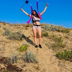 May include: A woman in a pink and white patterned bikini top and matching shorts is holding two blue and purple streamers with black handles. She is standing on a sandy dune with a blue sky in the background.