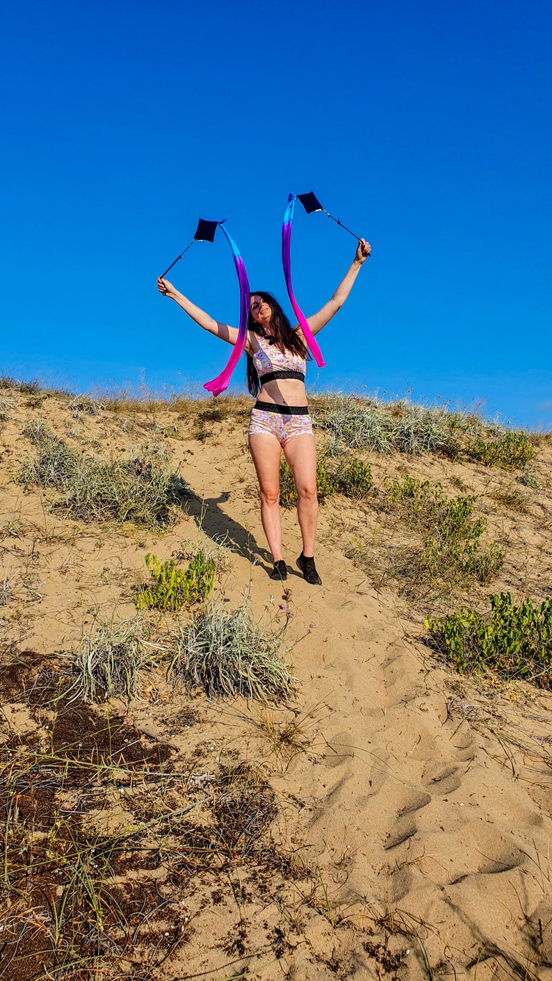 May include: A woman in a pink and white patterned bikini top and matching shorts is holding two blue and purple streamers with black handles. She is standing on a sandy dune with a blue sky in the background.