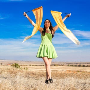 May include: A person in a green dress stands in a field, holding two orange and white flags against a bright blue sky. The dress is short and flared, and the flags are flowing in the wind. The scene is set in a rural landscape.