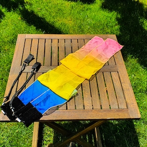 May include: Two colourful fabric streamers with black straps. The streamers are folded and laid on a wooden table. The streamers are pink, yellow, and blue. The table is outdoors on a grassy surface.