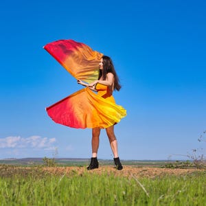 Könnte beinhalten: Eine Person in einem orangefarbenen Kleid hält einen fließenden Stoff in Rot-, Orange- und Gelbtönen vor einem strahlend blauen Himmel. Die Person trägt schwarze Stiefel und steht auf einer Wiese.