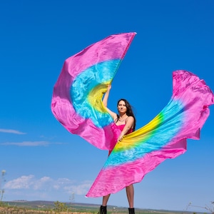 May include: A woman in a pink dress holds two large, colorful fabric flags. The flags are pink, blue, yellow, and green. The woman is standing in a field of green grass with a blue sky in the background.