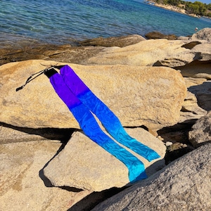 Peut inclure: Longs rubans flottants dans un dégradé de couleurs allant du violet au bleu et turquoise, avec des attaches noires. Les rubans sont drapés sur de gros rochers clairs près de la mer. La scène se déroule par une journée ensoleillée.