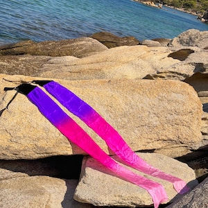 May include: Two long, flowing silk ribbons with a color gradient from purple to pink. The ribbons are lying on a large, light-colored rock formation near the ocean. The ribbons have black handles.