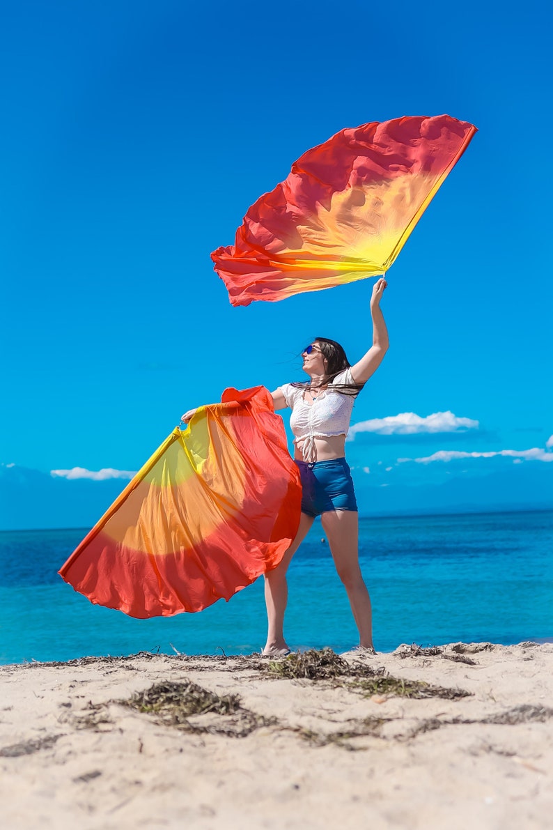 May include: A person on a sandy beach holds two large, colorful flags against a bright blue sky. The flags transition from red to yellow. The person wears a white top and denim shorts, with sunglasses.