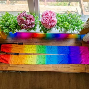 May include: Two long, rectangular fabric streamers featuring a vibrant rainbow gradient pattern that transitions from red to orange, yellow, green, blue, and purple. Each streamer has a black fabric handle attached to one end with a black strap.