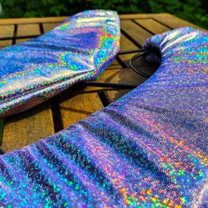 Könnte beinhalten: Ein Paar geschwungene, halbmondförmige Stoffüberzüge mit einer lebendigen, holografischen, lichtreflektierenden Oberfläche, die ein Spektrum schillernder Farben wie Violett, Blau und Grün zeigt und eine texturierte, schimmernde Oberfläche aufweist.