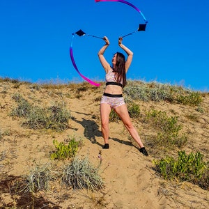 May include: A person in a pink and white patterned sports bra and shorts is twirling two ribbons in the air. The ribbons are pink, blue, and purple. The person is standing on a sandy hill with green plants in the background. The sky is blue.