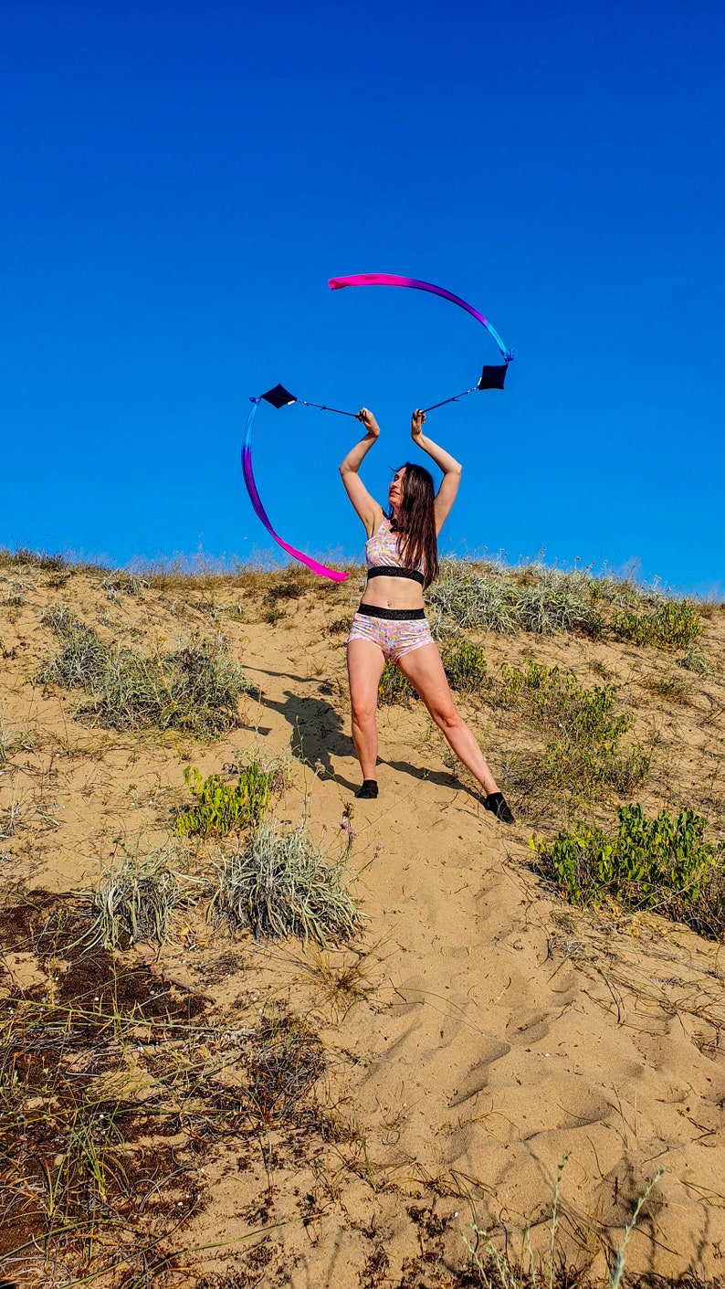 May include: A person in a pink and white patterned sports bra and shorts is twirling two ribbons in the air. The ribbons are pink, blue, and purple. The person is standing on a sandy hill with green plants in the background. The sky is blue.