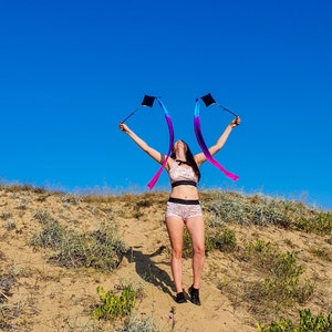 May include: A person wearing a floral patterned sports bra and matching shorts is holding two rainbow ribbon wands in the air. The person is standing on a sandy dune with green grass and blue sky in the background.