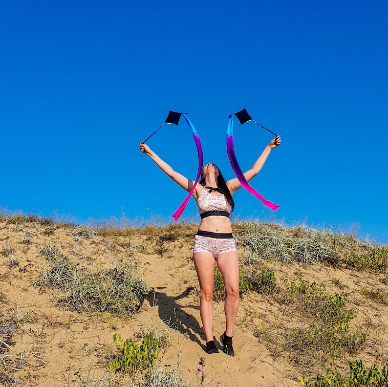 May include: A person wearing a floral patterned sports bra and matching shorts is holding two rainbow ribbon wands in the air. The person is standing on a sandy dune with green grass and blue sky in the background.