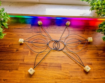May include: A black metal fire prop featuring two interconnected circular handles and four symmetrical wing-like frames. Each frame extends outward to a square-shaped, off-white Kevlar wick secured with a silver metal screw at the end of each arm.