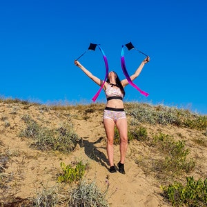 May include: A person wearing a pink and white patterned sports bra and matching shorts is holding two blue, pink, and purple ribbon streamers in the air. The person is standing on a sandy hill with green plants in the background. The sky is blue.