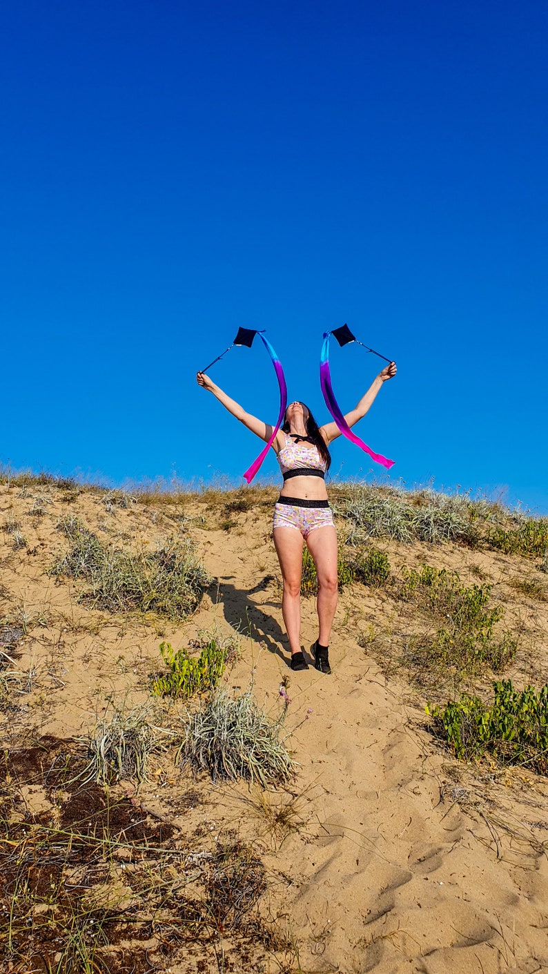 May include: A person wearing a pink and white patterned sports bra and matching shorts is holding two blue, pink, and purple ribbon streamers in the air. The person is standing on a sandy hill with green plants in the background. The sky is blue.