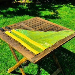 Puede incluir: Una bolsa de malla amarilla y verde con correas negras, doblada y colocada sobre una mesa de madera. La mesa está al aire libre en un césped.