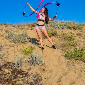 May include: A woman in a pink and blue patterned sports bra and matching shorts is twirling a long, colorful ribbon in a sandy, grassy area. The sky is blue.