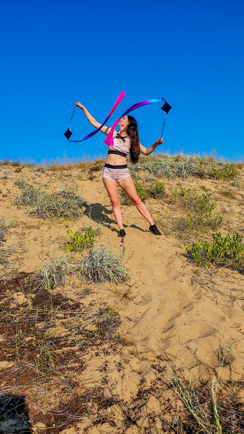 May include: A woman in a pink and blue patterned sports bra and matching shorts is twirling a long, colorful ribbon in a sandy, grassy area. The sky is blue.