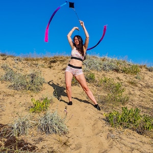 May include: A woman in a floral print sports bra and shorts is twirling a long, colorful ribbon in a sandy, grassy area. The ribbon is attached to a black handle.