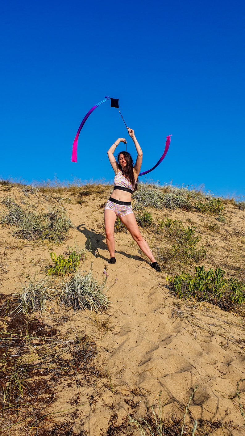 May include: A woman in a floral print sports bra and shorts is twirling a long, colorful ribbon in a sandy, grassy area. The ribbon is attached to a black handle.