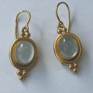 May include: A pair of gold-colored earrings. Each earring features an oval, pale blue gemstone set within a gold frame. The earrings have a hook design with small gold beads at the top and bottom of the gemstone setting.