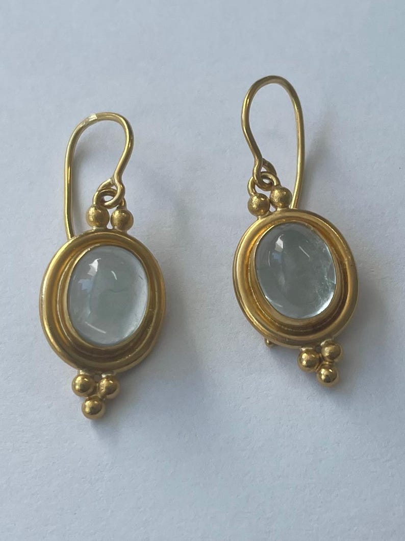 May include: A pair of gold-colored earrings. Each earring features an oval, pale blue gemstone set within a gold frame. The earrings have a hook design with small gold beads at the top and bottom of the gemstone setting.