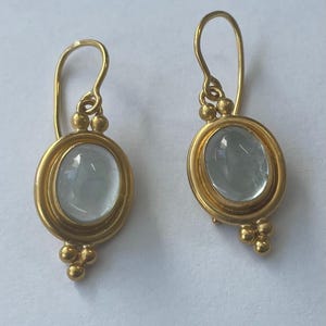 May include: A pair of gold-colored earrings with oval, light blue gemstones. The earrings have a simple design with a hook and small gold beads. The gemstones are set in a gold frame.