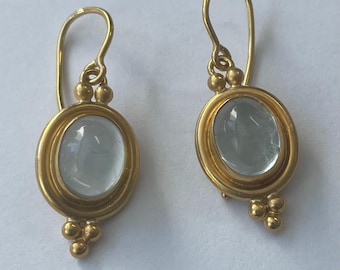 May include: A pair of gold-colored earrings with oval, light blue gemstones. The earrings have a simple design with a hook and small gold beads. The gemstones are set in a gold frame.