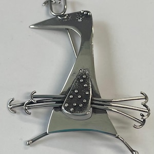 May include: A polished silver metal sculpture in an abstract bird-like form. The sculpture features a triangular body, curved legs, and a head with a U-shaped detail. Small, round accents adorn the body, and curved wire elements extend from the sides.