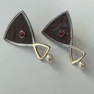 May include: A pair of triangular earrings featuring a dark, textured brown center framed in silver. Each earring has a small, round, deep red gemstone set in the middle and a dangling gold-toned triangular accent tipped with a white pearl.